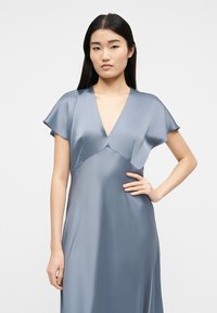 Γυναίκα με μακριά μαύρα μαλλιά που φοράει ένα ανοιχτό μπλε σατέν φόρεμα με κοντά μανίκια και V-λαιμόκοψη, στέκεται με το ένα χέρι στη μέση.