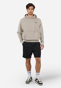 Hoodie beige con tasca a marsupio e logo, abbinato a pantaloni corti neri. Il modello indossa sneaker bianco e nere con calze bianche.