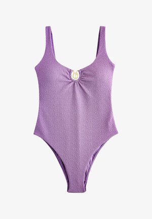 Femme en maillot de bain lavande texturé, assise sur un coussin blanc contre un mur en pierre rustique, une main touchant ses cheveux.