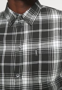 Camicia a scacchi bianco e nero con colletto abbottonato, realizzata in cotone, dotata di bottoni neri e un'etichetta sul lato anteriore.