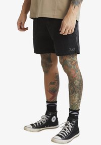 Shorts en velours côtelé noirs avec un logo brodé, portés avec des baskets montantes noires et des chaussettes côtelées. Des tatouages décorent les deux jambes.
