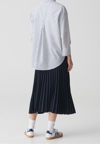 Personne portant une chemise blanche à fines rayures à manches longues, une jupe midi plissée noire, des chaussettes hautes blanches et des baskets blanches avec des rayures bleu marine.