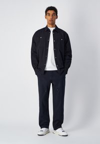 Marineblauwe shirtjack met twee borstzakken, gecombineerd met losse marineblauwe broek. Witte T-shirt eronder en witte sneakers met zwarte accenten.