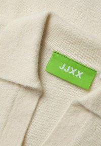 Maglione a maglia color crema con una consistenza morbida e un colletto ripiegato. Presenta un'etichetta verde brillante con la scritta "JJXX."