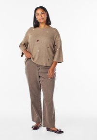 Maglione lavorato a maglia beige con maniche corte, caratterizzato da piccoli motivi a cuore rossi e bianchi. Abbinato a jeans marroni a zampa d'elefante e scarpe scure.