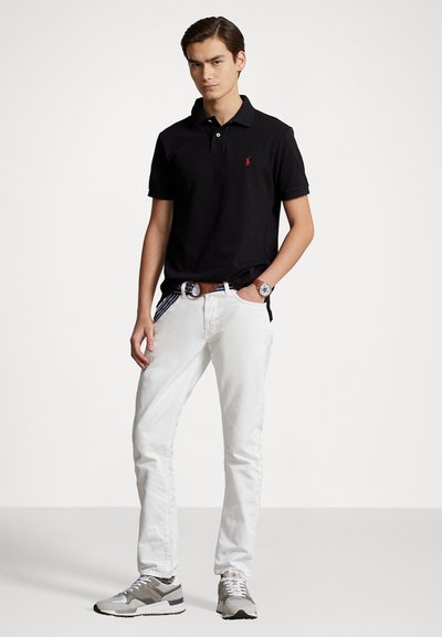 Svart poloskjorte med korte ermer og rødt logo, kombinert med hvite denimbukser og grå joggesko, med et teksturert brunt belte.