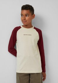 Crème kleurige t-shirt met bordeauxrode raglan mouwen. Heeft kleine zwarte tekst "SLVR STUDIOS | DEPARTMENT" aan de voorkant. Zachte katoenen stof.