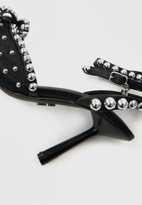 Melni augstpapēžu sandales ar metāliska sudraba kniedēm, ar teksturētu audumu un gludiem ādas akcentiem. Iekļauts regulējams potītes siksniņa.