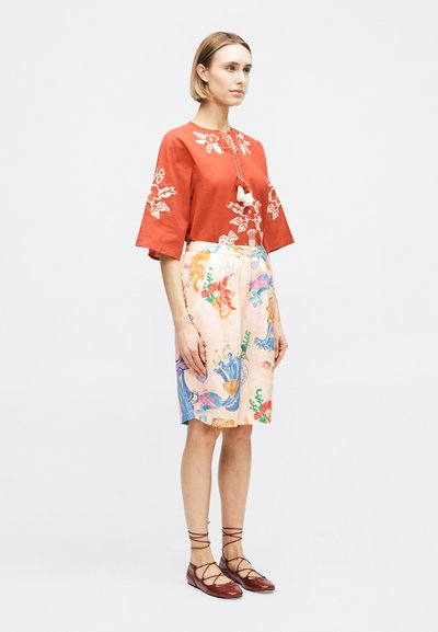 Femme debout portant un haut brodé couleur rouille avec des pompons et une jupe longueur genou avec des motifs abstraits colorés, ainsi que des chaussures plates marron à lacets.