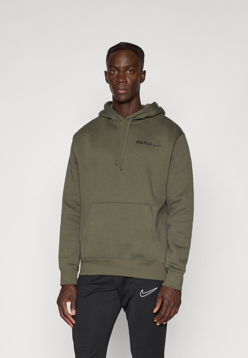 Felpa verde oliva con tasca frontale, cappuccio con coulisse e testo "PARIGI" in nero. Abbinata a pantaloni neri con logo Nike bianco.