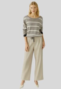 Vrouw in een grijs-wit gestreept truitje, beige wijde broek met een drawstring taille en witte puntige hakken, staande.