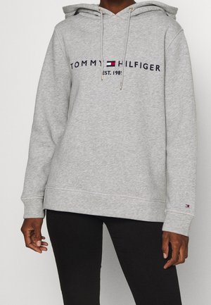 Person iført en lysegrå Tommy Hilfiger-hættetrøje med logo og snore, kombineret med sorte bukser, stående mod en ensfarvet baggrund.
