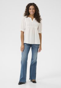 Blusa bianca con yoke testurizzato e maniche corte a sbuffo, abbinata a jeans blu a campana e ballerine nere lucide. Design semplice e pulito.