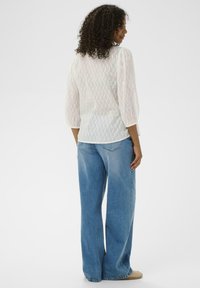 Femme aux cheveux bouclés portant un chemisier blanc texturé à manches 3/4 et un jean bleu ample, se tenant de dos sur un fond blanc.