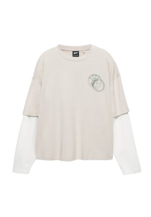 Beige T-shirt med korte ærmer lagdelt over en hvid langærmet skjorte. Har et lille grønt grafiktryk på brystområdet.