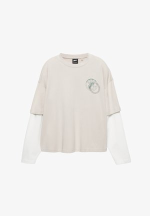 T-shirt beige à manches courtes porté par-dessus une chemise blanche à manches longues. Présente un petit imprimé graphique vert sur la poitrine.
