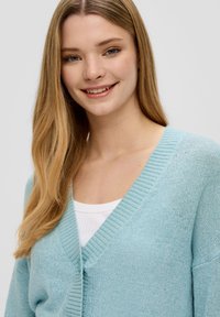 Jasnoniebieski kardigan z dekoltem w serek i prążkowanymi mankietami. Miękka teksturowana tkanina, z zapięciem na guziki z przodu, nałożony na białą bluzkę.