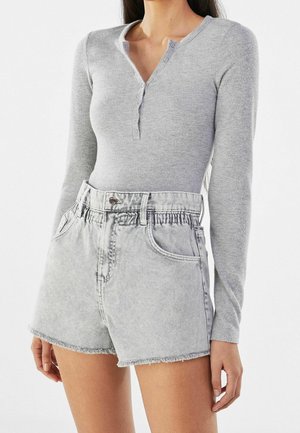 Kobieta ubrana w dopasowaną jasnoszarą bluzkę henley z długim rękawem, włożoną do jasnych, wysokich szortów jeansowych z talią paperbag i postrzępionymi brzegami.