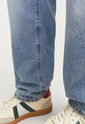 Henkilö, joka pukeutunut sinisiin farkkuihin ja valkoisiin lenkkareihin, joissa on vihreät raidat ja kuminahkaiset pohjat, seisoo valkoisella pinnalla.