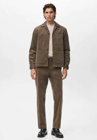 Chaqueta de pana marrón con bolsillos en la parte delantera, combinada con pantalones a juego, un suéter ligero debajo y zapatos negros. Diseño minimalista.