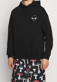 Schwarzer Kapuzenpullover mit Fronttasche, der ein kleines, farbenfrohes Grafikdesign auf der Brust trägt. Das Material wirkt weich und lässig.