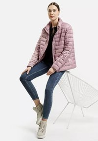 Jasnoróżowa pikowana kurtka z kołnierzem, noszona na czarnej bluzce, w połączeniu z niebieskimi obcisłymi dżinsami i beżowymi sneakersami. Biała krzesło w tle.