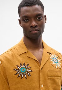 Κοντομάνικο πουκάμισο σε μουστάρδι χρώμα από λινάρι με κεντητά μοτίβα ήλιου σε καφέ, λευκό και μπλέ. Διαθέτει τσέπη στο στήθος και ραφές ως διακοσμητικά στοιχεία.