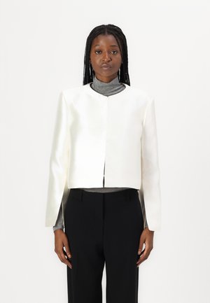 Giacca cropped in satin bianco con maniche lunghe, scollatura rotonda e apertura frontale. Indossata sopra un dolcevita grigio, abbinata a pantaloni neri.