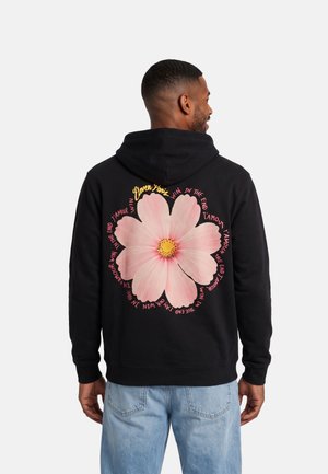 Un hombre lleva una sudadera negra con capucha y unos vaqueros azul claro; en la espalda de la sudadera hay una gran flor rosa y un texto circular.