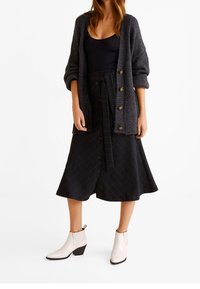 Grauer gestrickter Cardigan mit großen Knöpfen, schwarze mit Knöpfen versehene Röhre mit Rautenmuster und weiße Knöchelstiefel mit Blockabsatz.