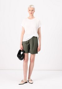 Haut blanc à manches courtes en rib et shorts verts olives jusqu'aux genoux. Tient un sac à main noir, porte des chaussures beiges à brides.