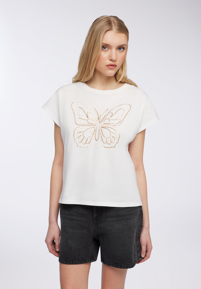 T-shirt bianca a maniche corte con un grande design di farfalla dorata, abbinata a pantaloni corti in denim neri. Il tessuto sembra morbido e leggero.