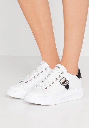 Białe niskie sneakersy z czarnymi akcentami na pięcie i logo postaci z kreskówki po bokach, noszone na stopach na neutralnym tle.