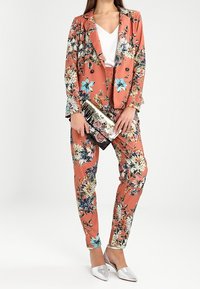 Floralanzug in Koralle mit einem doppelt knöpfbaren Blazer und engen Hosen. Ergänzt durch ein metallisches Clutch und silberne Ballerinas.