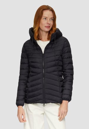 Veste rembourrée noire avec fermeture éclair sur le devant, design matelassé en chevron, capuche réglable avec cordon de serrage et poignets côtelés. Le modèle la porte par-dessus un pull blanc.