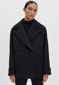 Cappotto doppiopetto nero con ampi risvolti, tessuto testurizzato e due tasche frontali, con grandi bottoni neri.