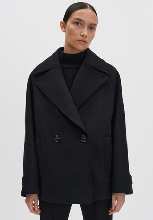 Cappotto doppiopetto nero con ampi risvolti, tessuto testurizzato e due tasche frontali, con grandi bottoni neri.