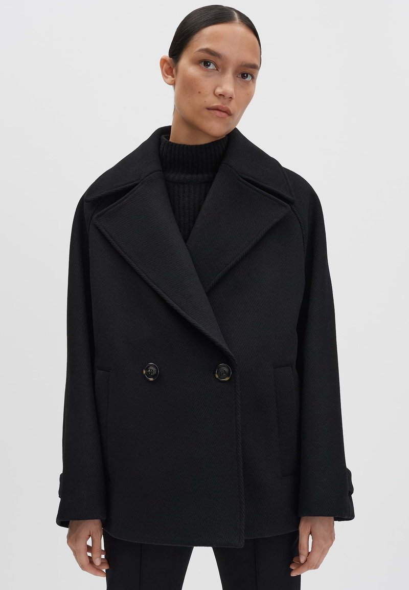 Cappotto doppiopetto nero con ampi risvolti, tessuto testurizzato e due tasche frontali, con grandi bottoni neri.