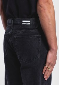 Los jeans de mezclilla negra presentan una etiqueta con "DRDENIM" en la cinturilla. Los bolsillos traseros están cosidos con un diseño clásico. Textura suave.