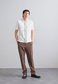 Weißes Kurzarmhemd mit schwarzem Besatz, kombiniert mit braunen Hosen. Das Model steht auf einem grauen Boden und trägt dunkle Schuhe mit weißen Schnürsenkeln.