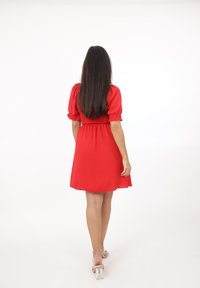 Vrouw met lang donker haar in een rode knielange jurk en doorzichtige hakken die wegloopt op een witte achtergrond.