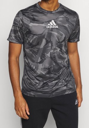Mies, jolla on musta ja harmaa maastokuvioinen Adidas-t-paita ja mustat Adidas-housut, seisoo yksiväristä taustaa vasten.