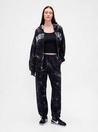 Mulher em calças de jogging pretas com padrão tie-dye, um casaco de zip com capuz combinando e letras brancas, top cropped preto e tênis Nike pretos.