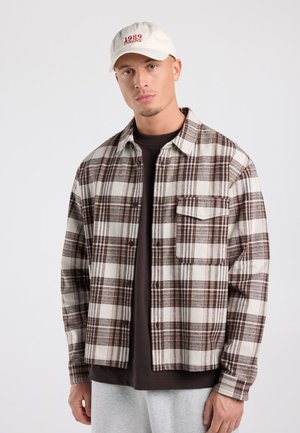 Jonge man die een wit-bruin geruit overhemd draagt, een zwart ondershirt, grijze broek en een witte pet met rode tekst.