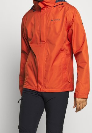 Mann trägt eine leuchtend orange Kapuzenjacke mit Fronttaschen und schwarze Hose, steht vor hellem, einfarbigem Hintergrund.