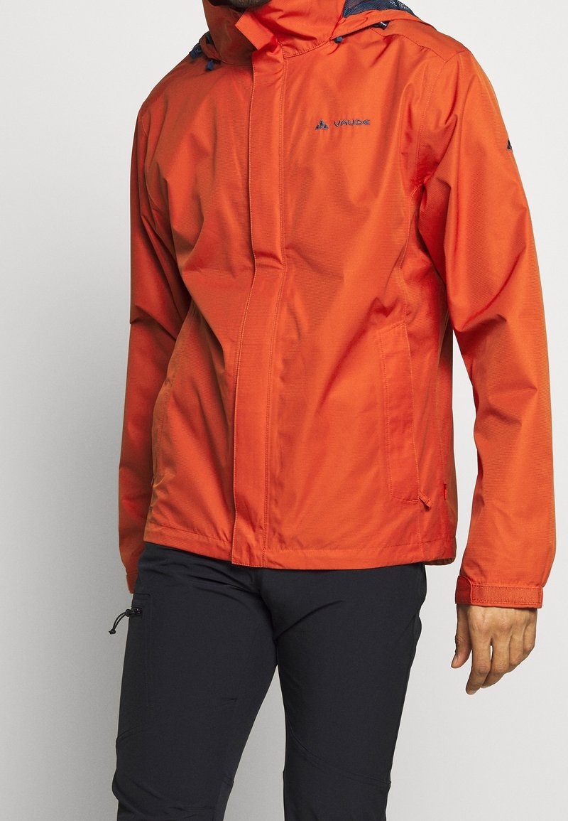 Homme portant une veste à capuche orange vif avec poches avant et pantalon noir, debout devant un fond uni et clair.