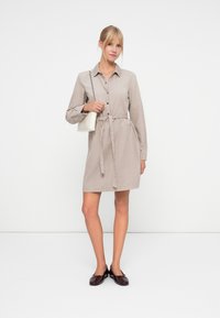 Button-up shirt dress in light beige fabric with a tie belt, long sleeves, and pockets. Paired with dark glossy loafers and a small white bag.