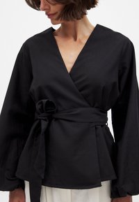 Blusa negra de manga larga con un profundo escote en V, mangas amplias y cintura atada. Fabricada en una tela suave con un corte suelto y fluido.