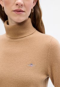 Sweter z golfem w kolorze kamelowym, wykonany z miękkiej dzianiny z prążkowanym kołnierzem; posiada małe haftowane logo na lewej piersi.