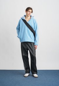 Un giovane si trova in piedi contro un muro bianco indossando un pile azzurro chiaro, pantaloni neri con strisce laterali, sneaker bianche e una borsa a tracolla nera.